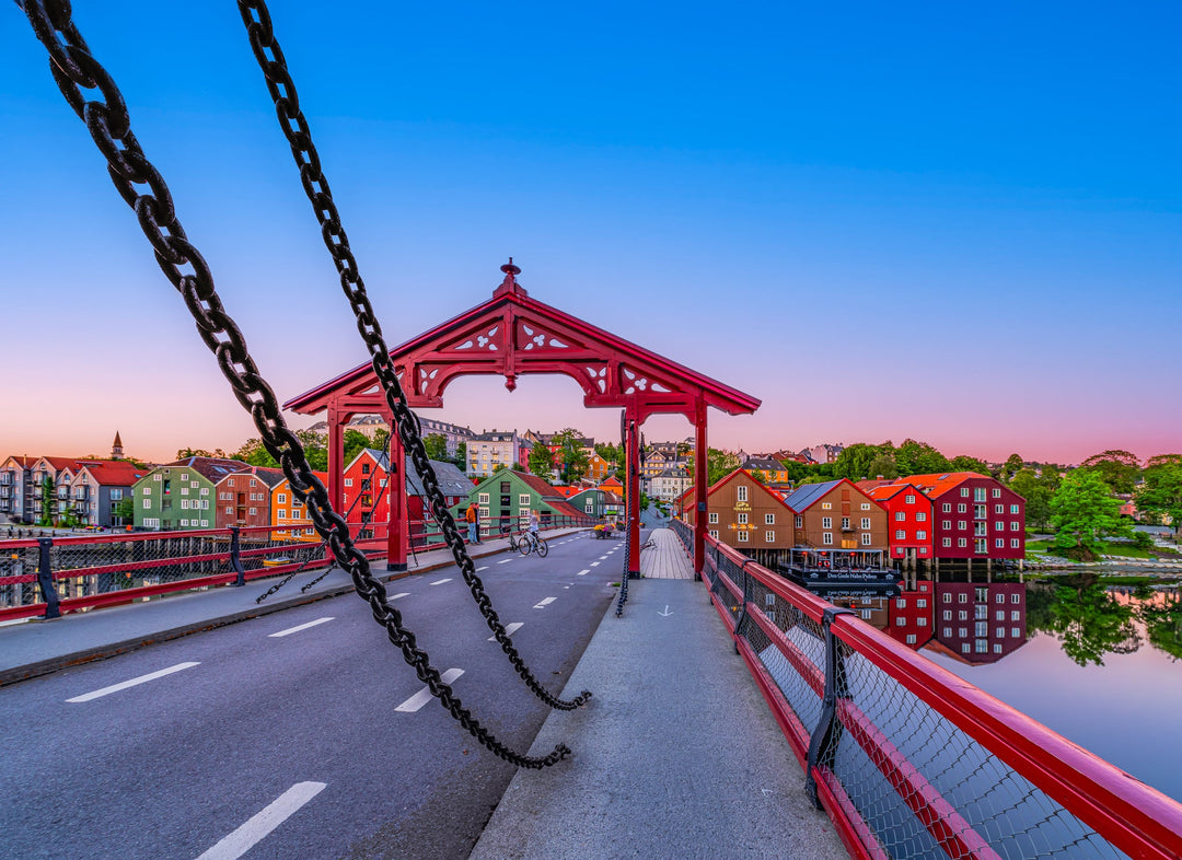 Gamle Bybro, a red wooden bridge in Trondheim, Norway, spans the Nidelva River with decorative gates and colorful wooden wharf buildings along the riverbanks, creating a scenic and historic view.